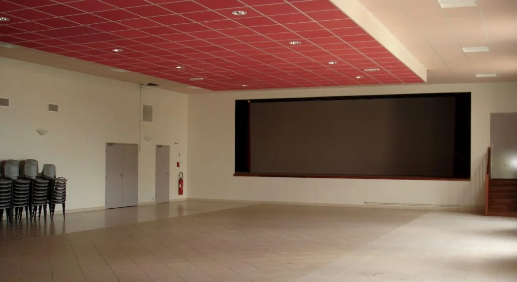 Intérieur de la salle polyvalente : une grande salle au plafond rouge, avec la scène au fond.